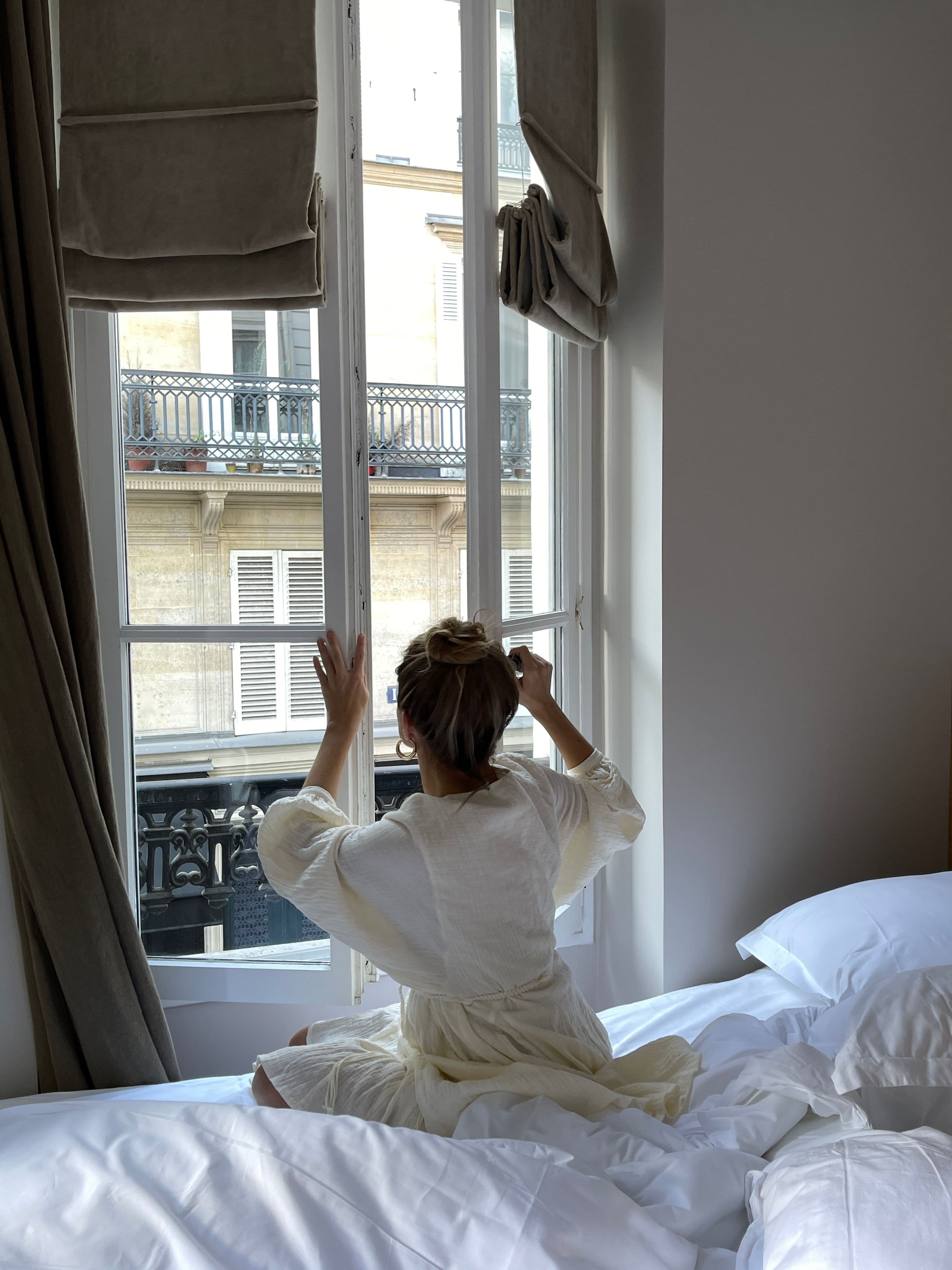 A woman in a linen dress opening tall shuttered windows onto a Parisian balcony, morning light filling the room.
